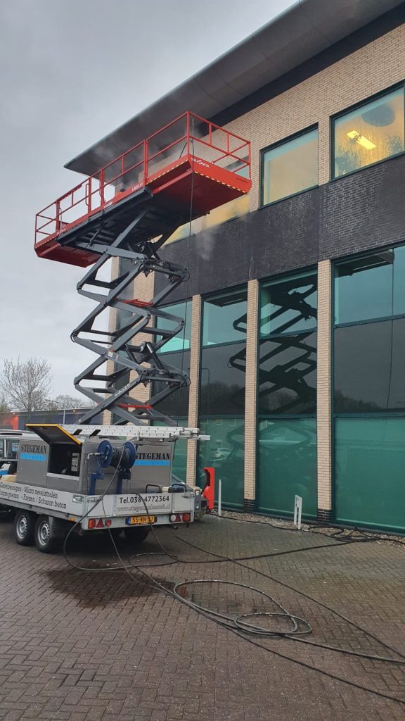 Schaarhoogwerker bij gevelonderhoud aan bedrijfspand met glazen gevel en grijze panelen