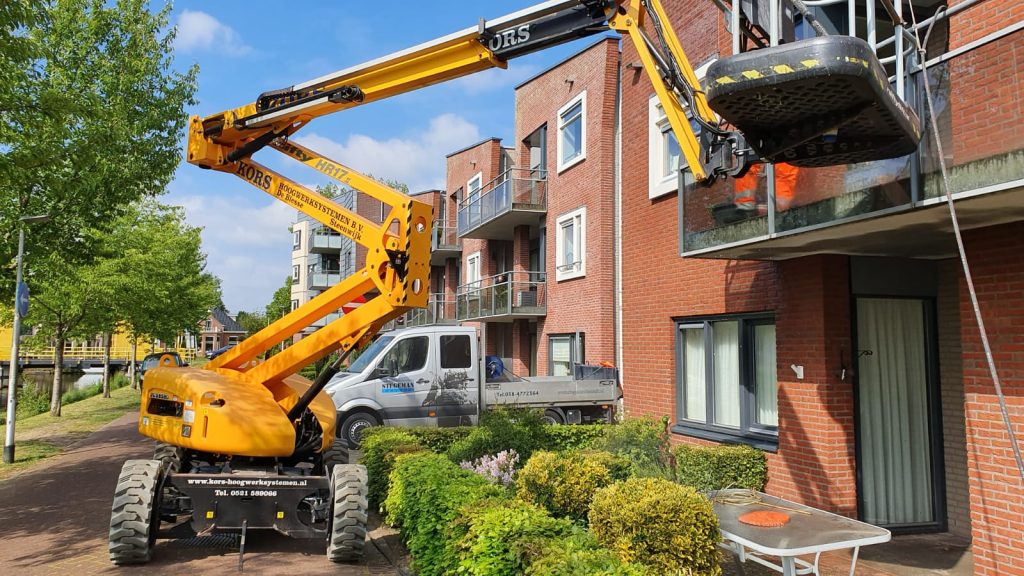 Gele hoogwerker met verhuislift verwijdert meubilair via balkon bij appartementencomplex