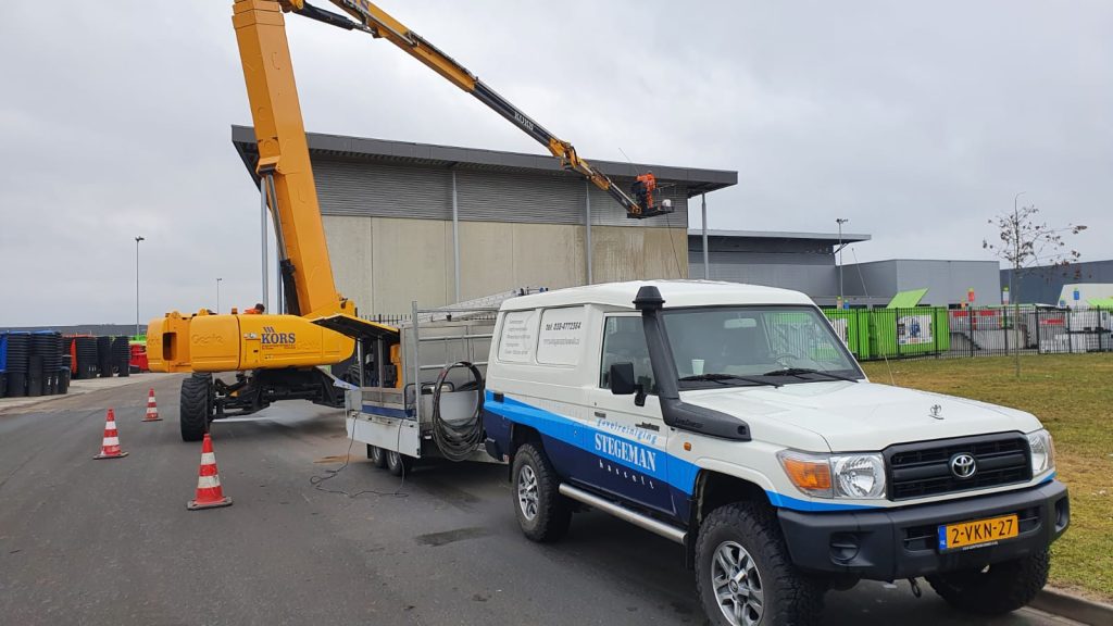 Hoogwerker met gestrekte arm naast servicevoertuig voor werkzaamheden aan gebouw met moderne gevelbekleding
